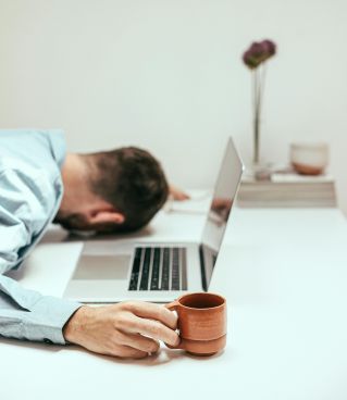 A fatigued man in an office setting asleep at his desk with a laptop and coffee mug.